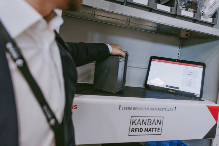 Eine Person stellt einen grauen Behälter in ein  Kanban RFID Regal. Im Hintergrund ist ein Monitor zu sehen, der Kanban Management Oberfläche zeigt. Oberhalb des Regals befinden sich weitere kleine Behälter mit Zubehör.